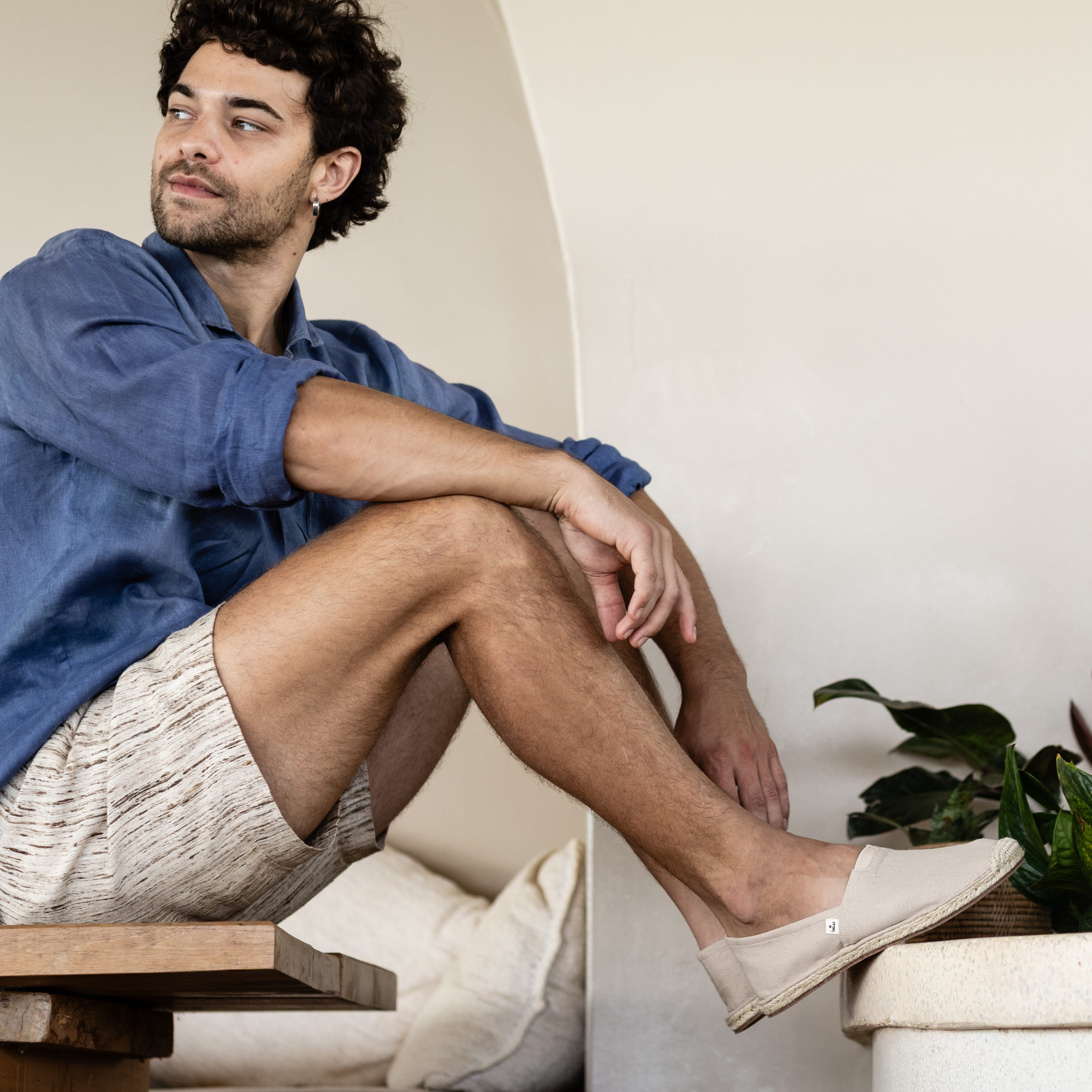 Man sitting on a bench wearing beige espadrille shoes, a blue shirt and shorts.