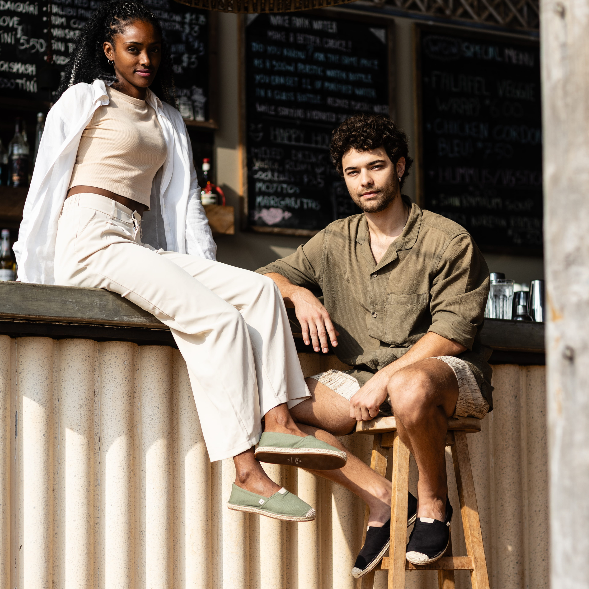 Two people sitting outdoors with a chalkboard menu in the background wearing summer espadrilles