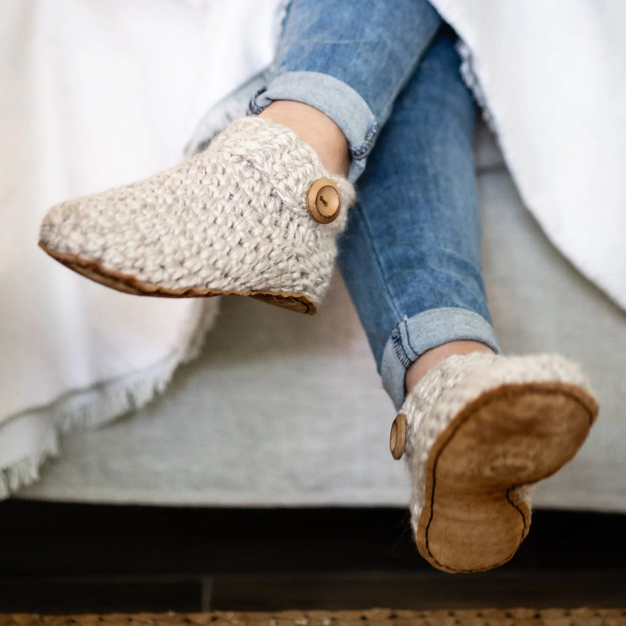 Woman wearing beige crocheted wool slippers with wooden buttons sat on a couch