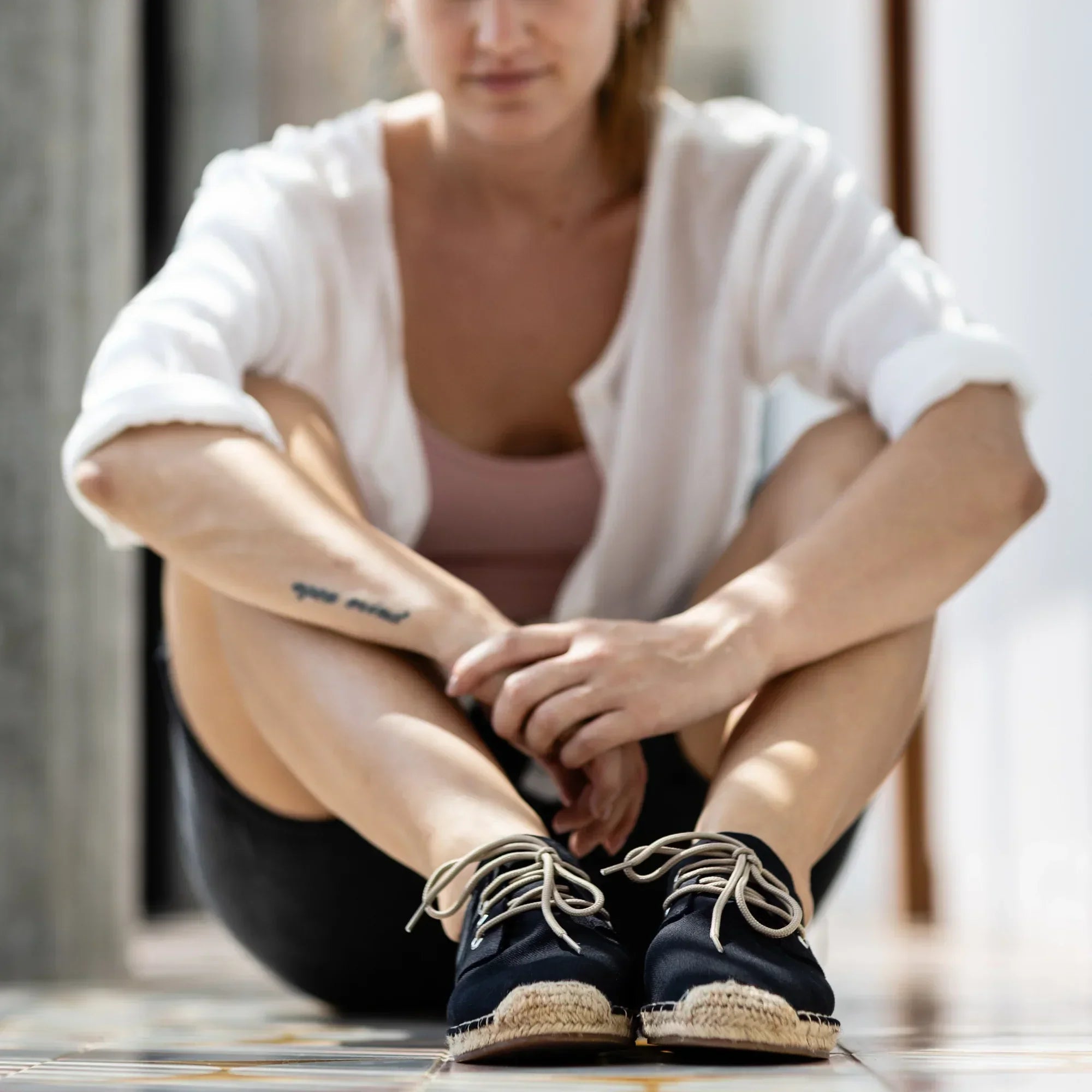 Woman sitting on a wooden floor wearing black lace-up espadrilles with jute rope soles.