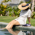 Woman sitting by a pool wearing a wide-brimmed hat and blue espadrilles.