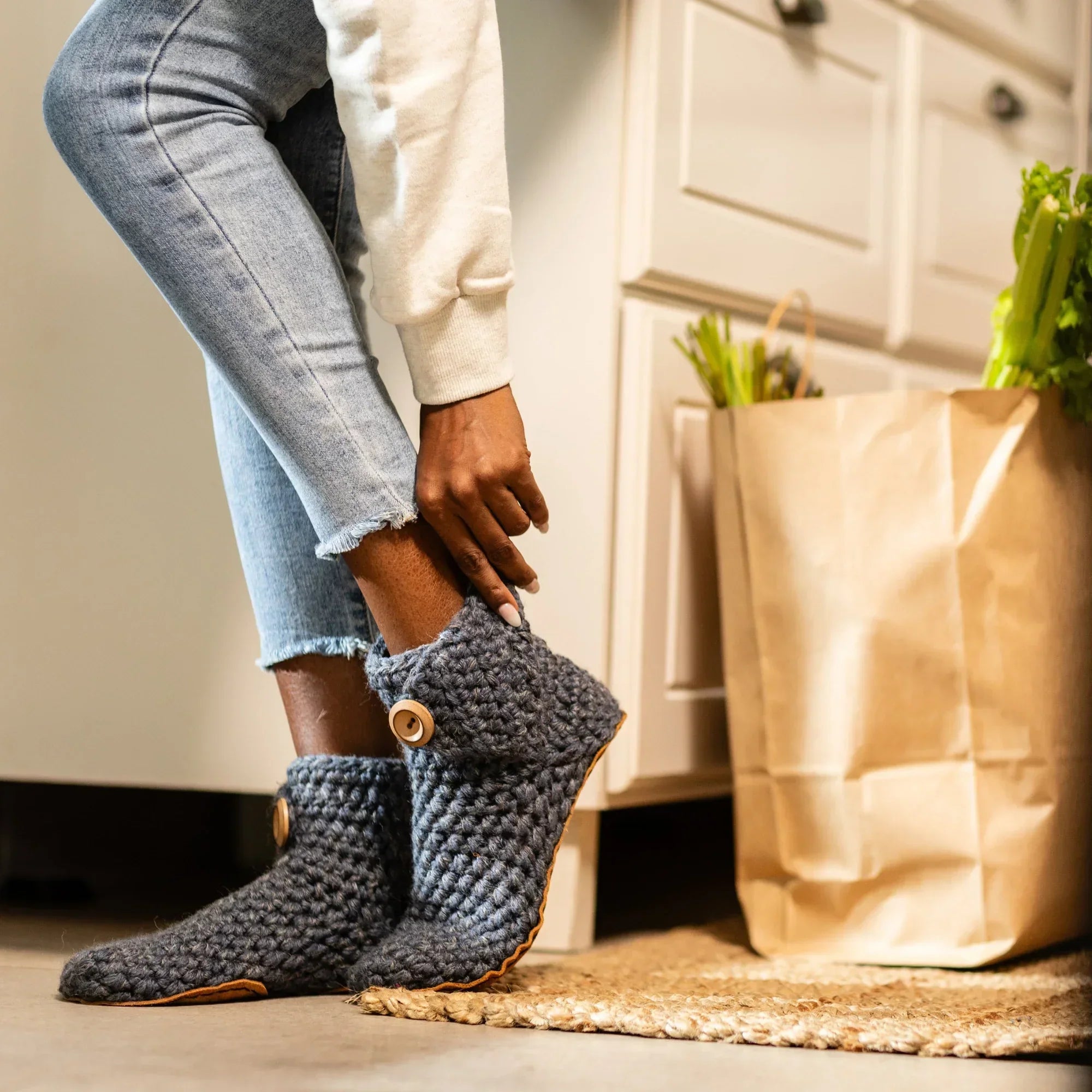 Person wearing blue knitted slipper boots with a brown paper bag and groceries in the background.