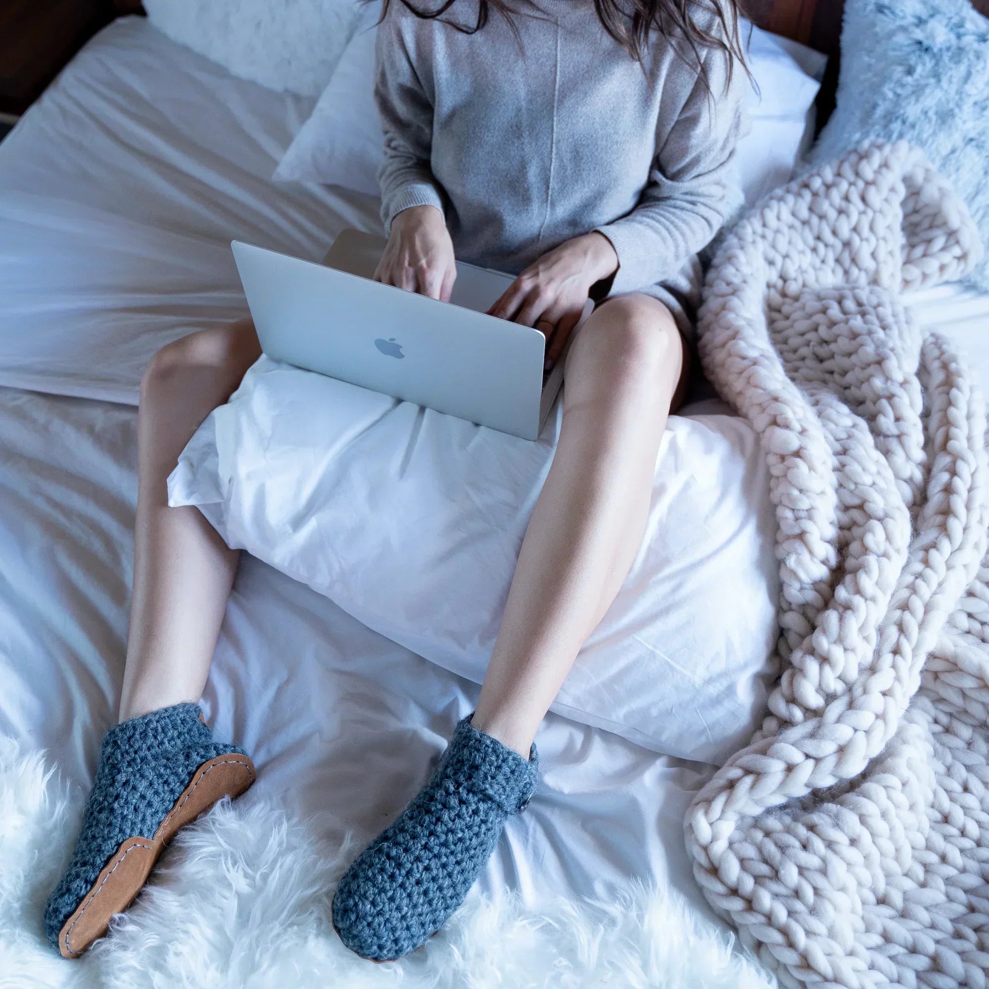 Woman using a laptop on a bed with cozy blankets and gray crocheted slippers by Kingdom of Wow.