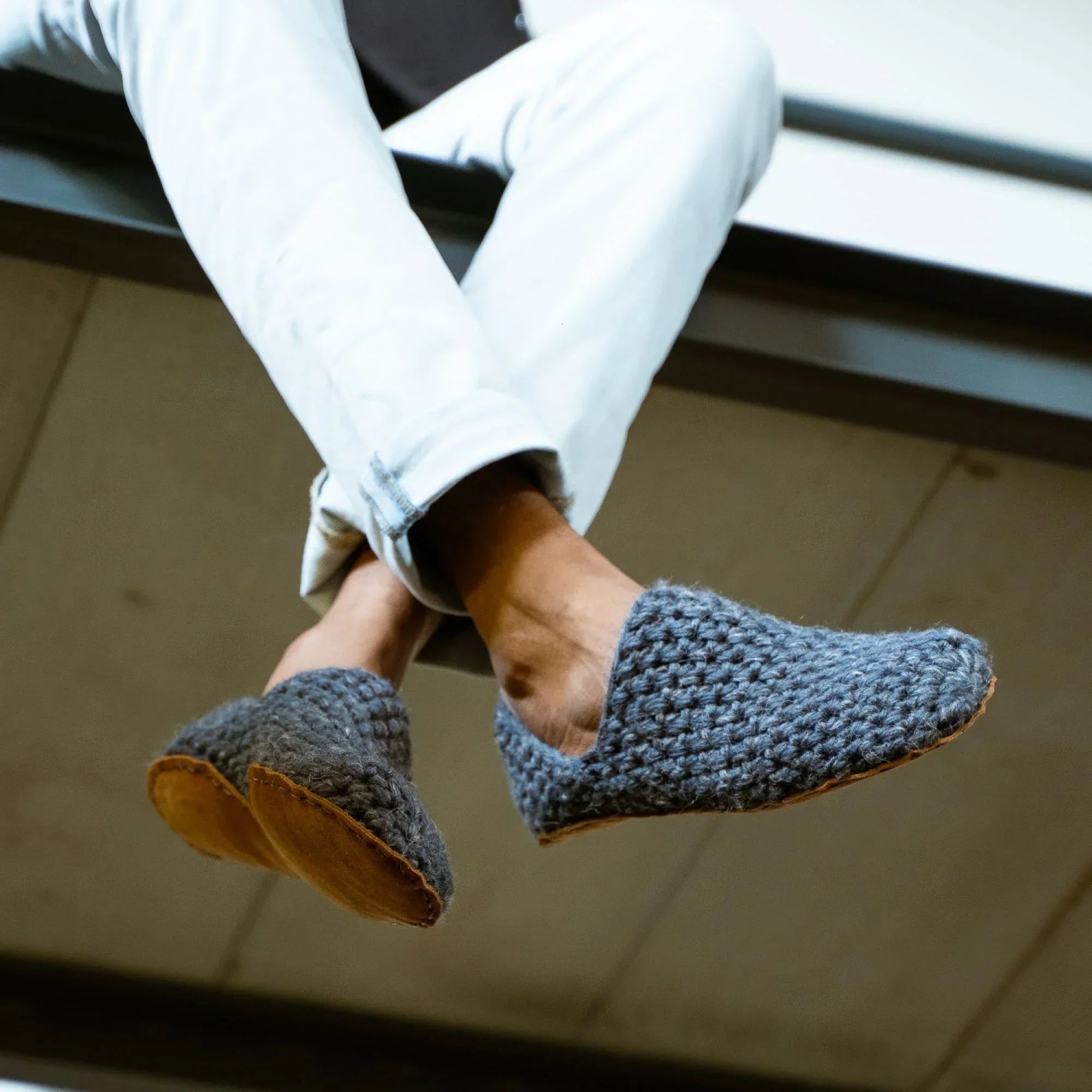 Person wearing gray crocheted wool slippers by Kingdom of Wow with denim pants sitting on a ledge