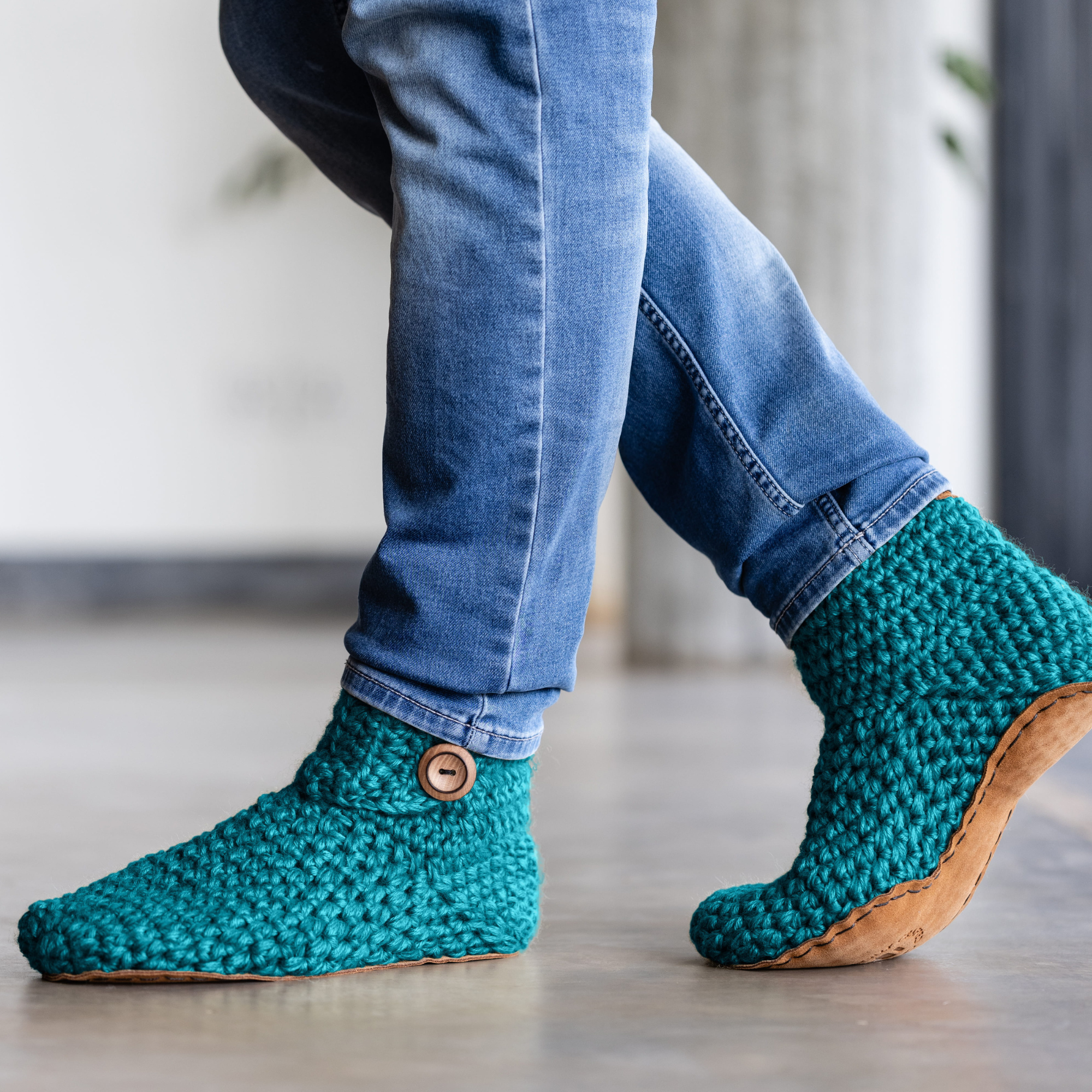 Teal knitted slippers with wooden buttons and leather sole worn by a person on a light polished cement floor.