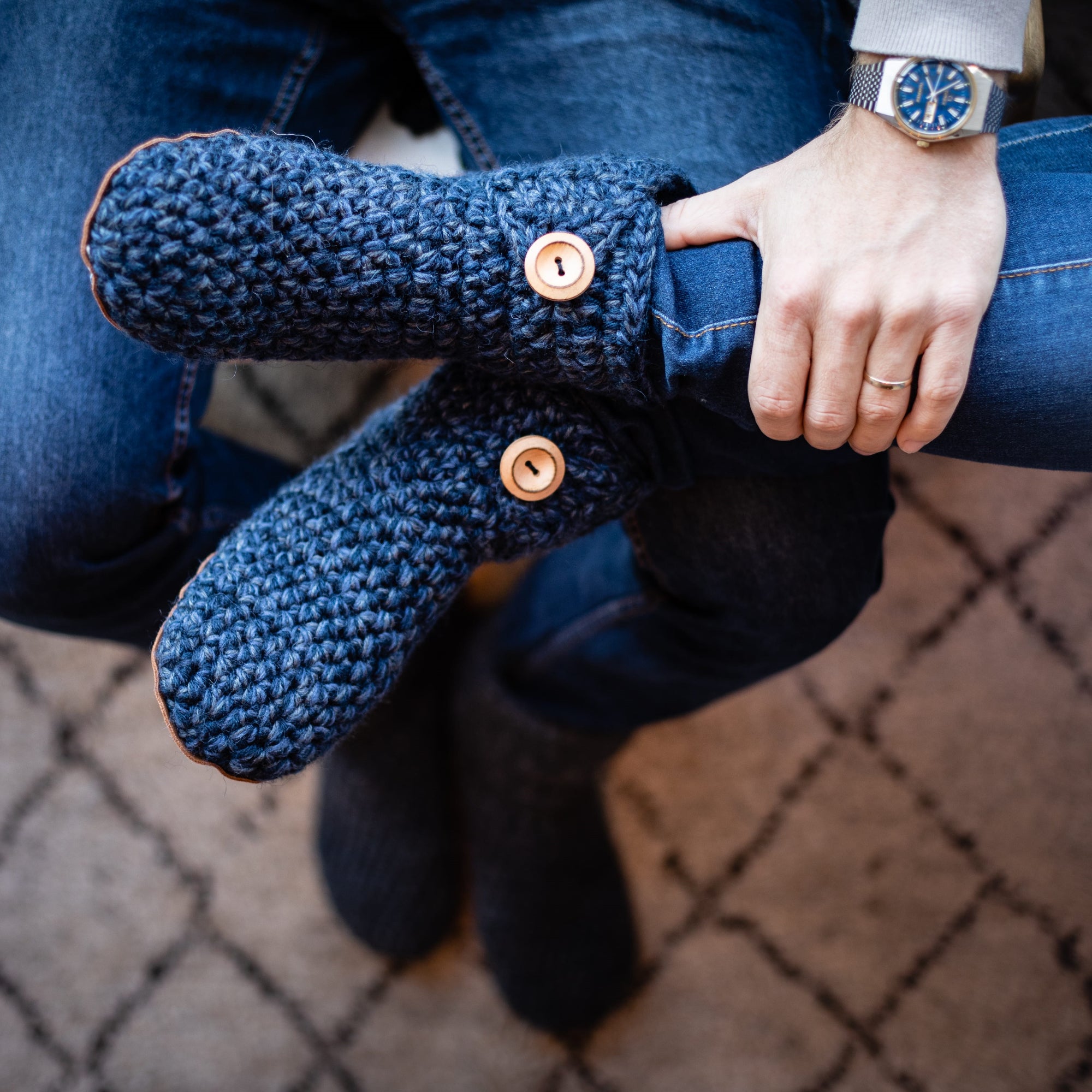 Person wearing blue knitted slippers with button details, with legs over husband's lap