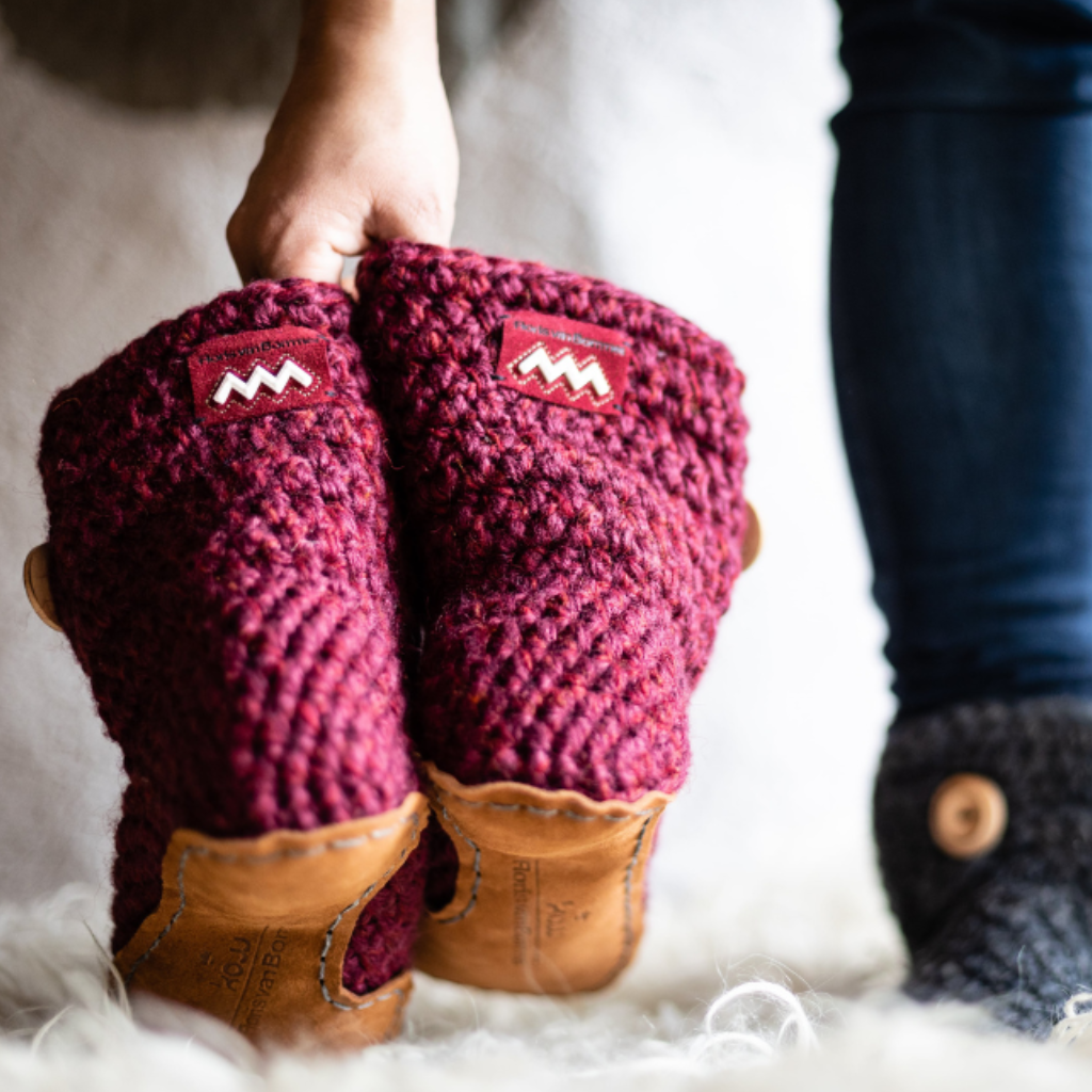 Person wearing maroon red crocheted slipper boots with brown soles on a soft surface.