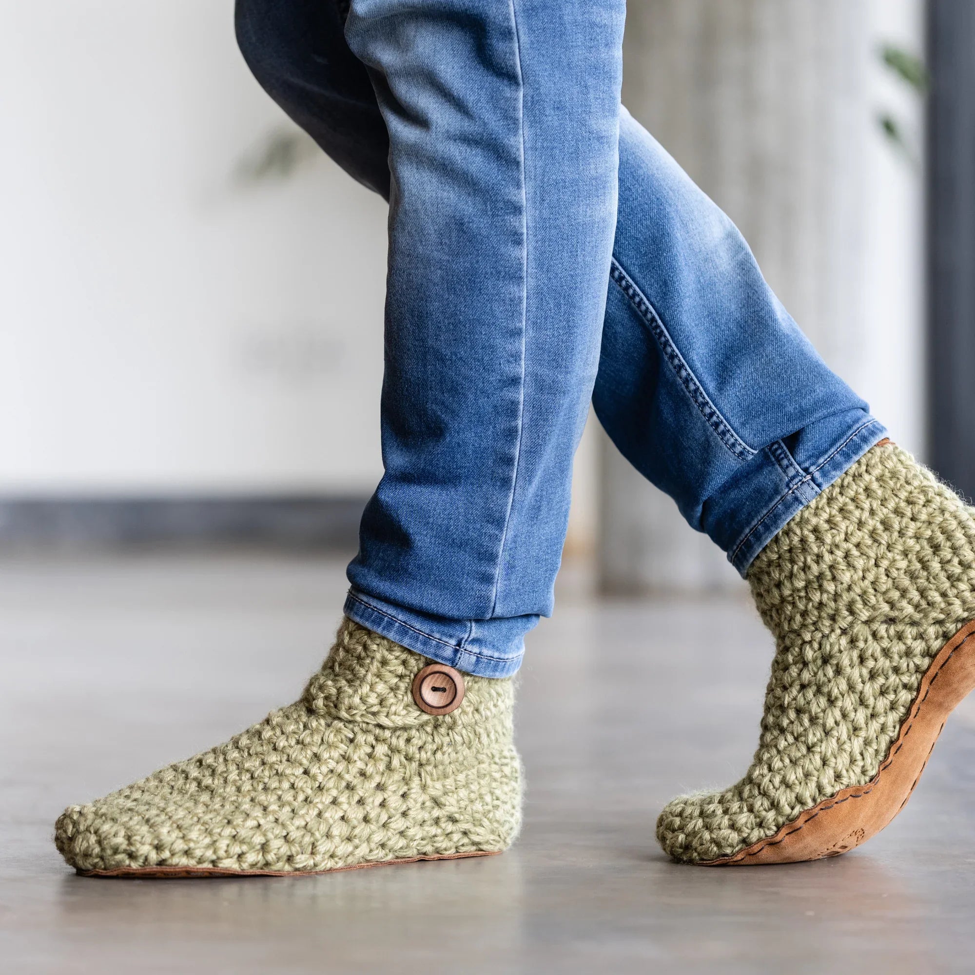 Green crocheted slipper boots with leather soles worn with blue jeans on a light wooden floor.