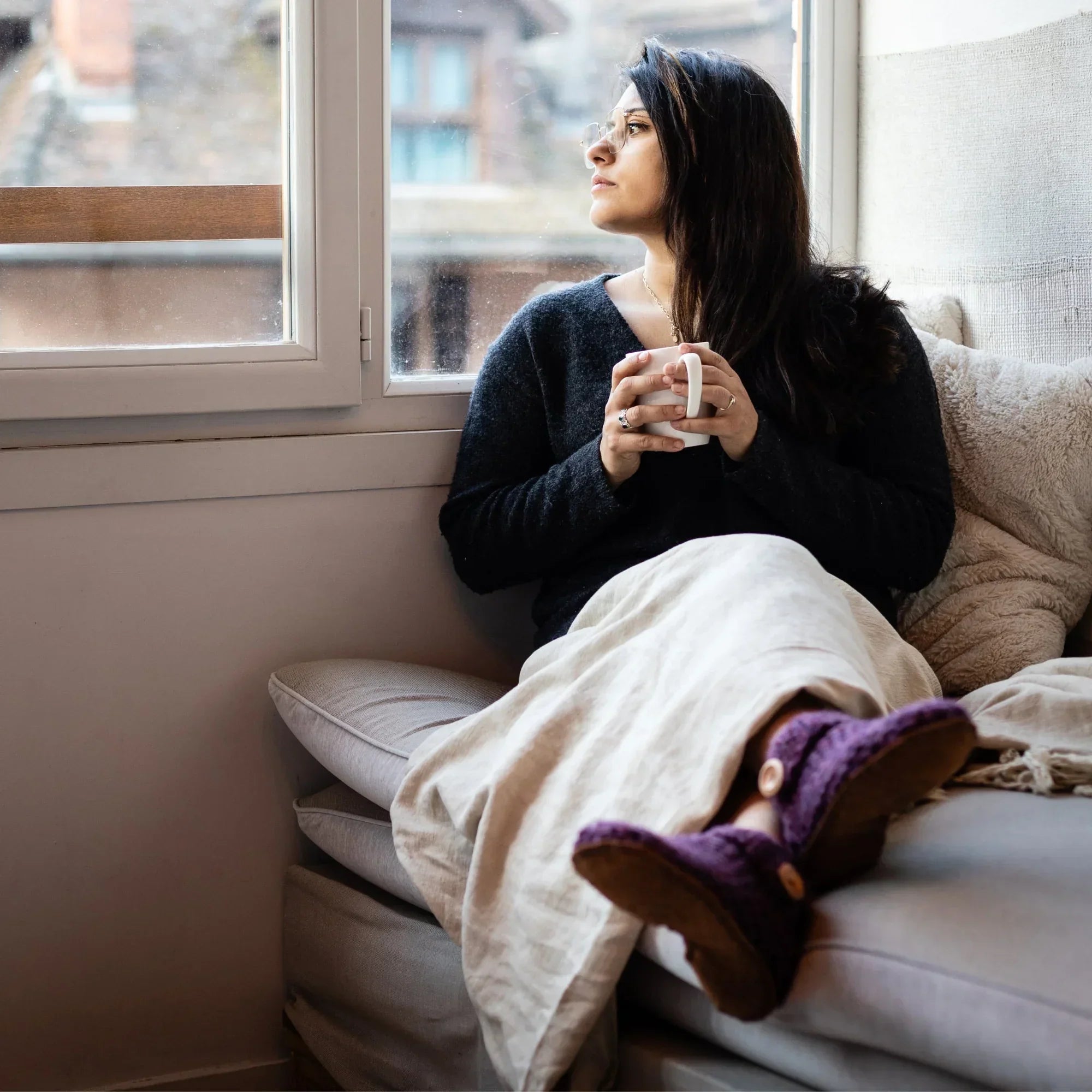 Woman sitting on a couch by a window, holding a mug, wrapped in a blanket with purple wool slippers.