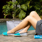 Woman wearing turquoise blue espadrilles by a poolside with a gray bag and tropical background