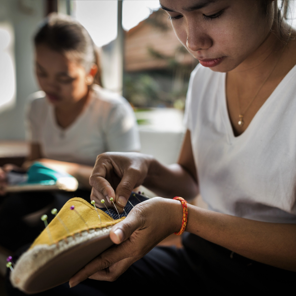 Woman sewing a yellow shoe with another person in the background.