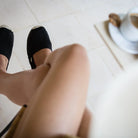 Woman wearing black espadrilles sitting on a white tiled floor with a blurred coffee cup and saucer in the background.
