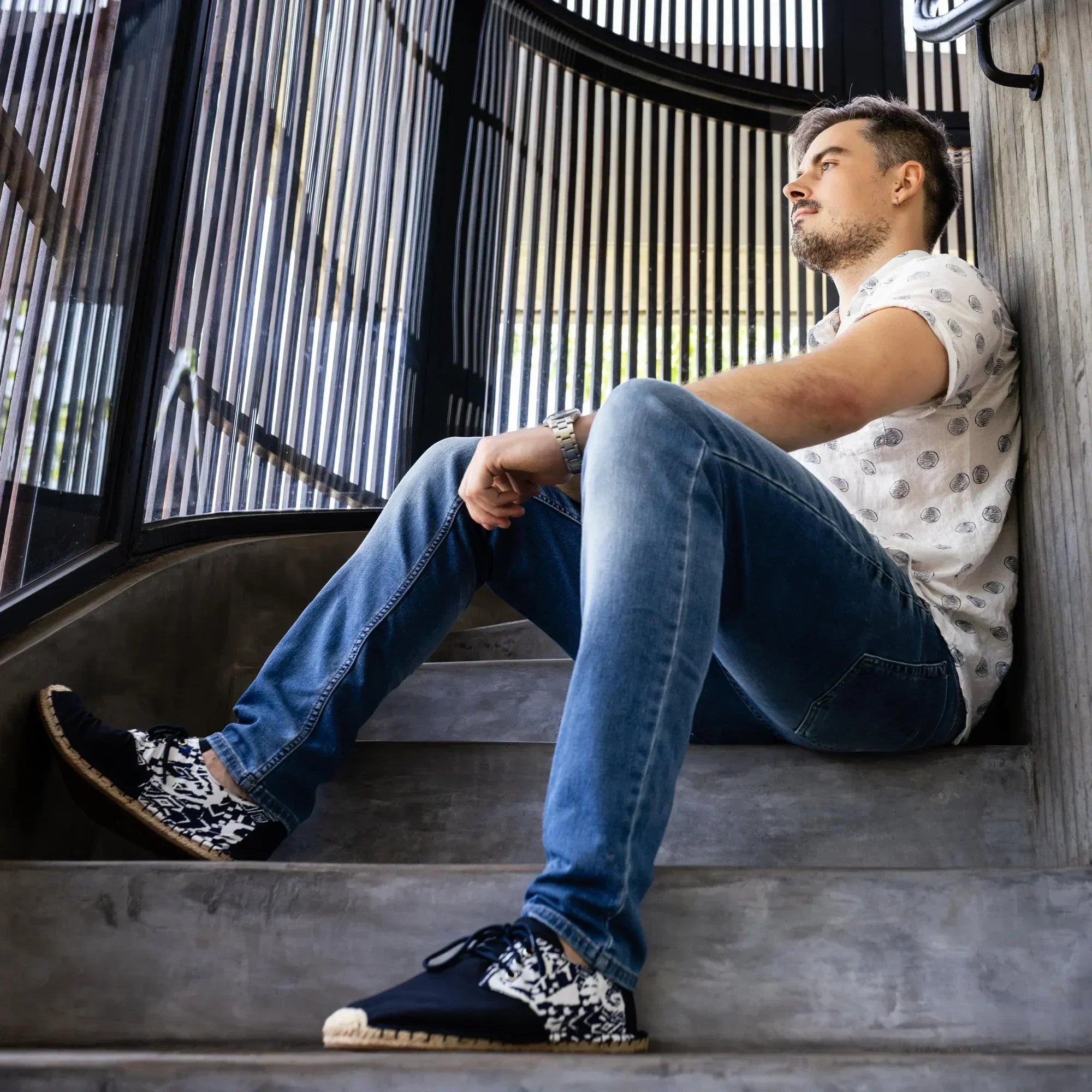 Man sitting on steps wearing blue jeans and dark blue espadrille sneakers, leaning against a wall.