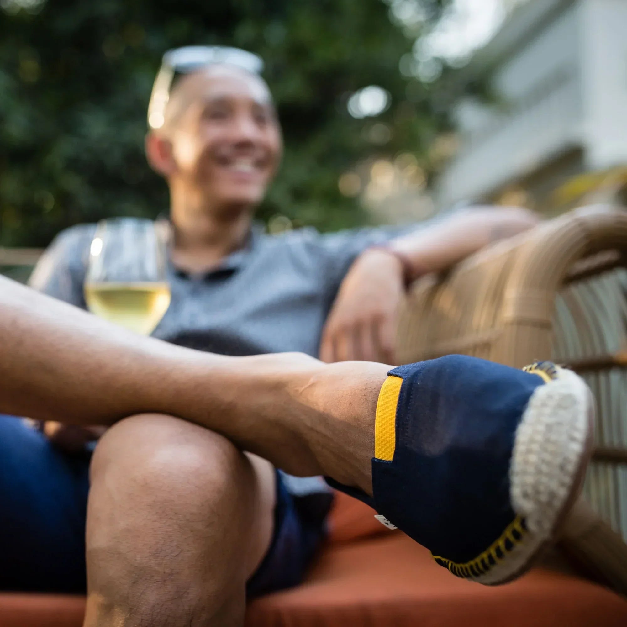 Man sitting outdoors with a glass of white wine, wearing dark blue espadrilles with yellow detailing.