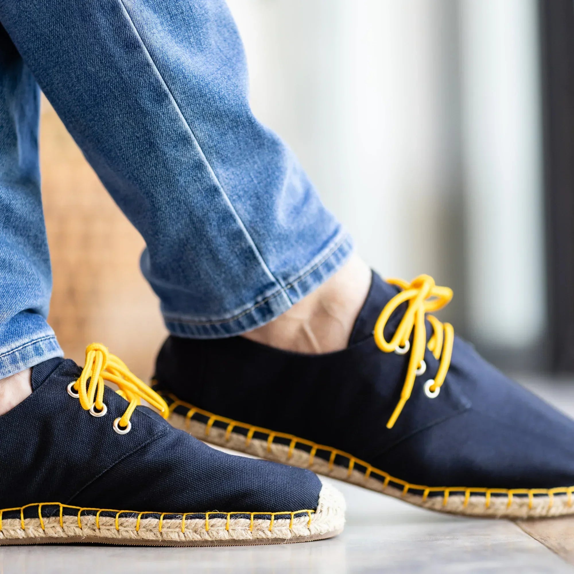 Navy blue espadrilles with yellow laces and stitching worn with blue jeans on a light wooden floor.