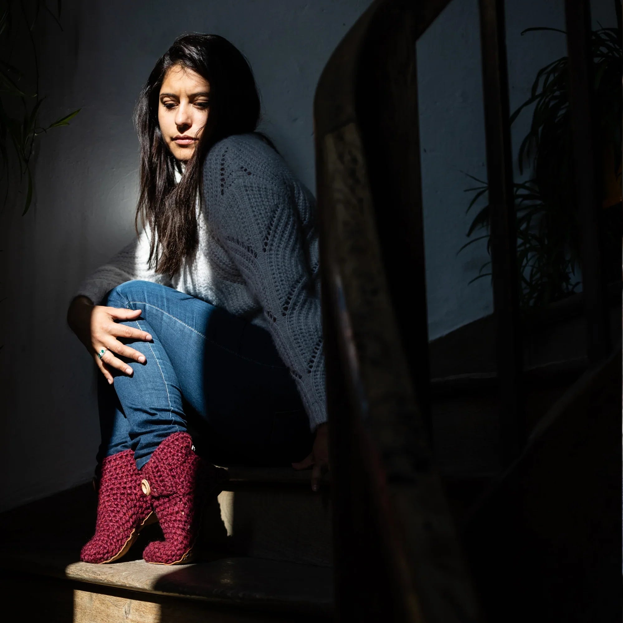 Woman sitting on a step wearing a white sweater, blue jeans, and red bootie wool slippers in a dimly lit setting.