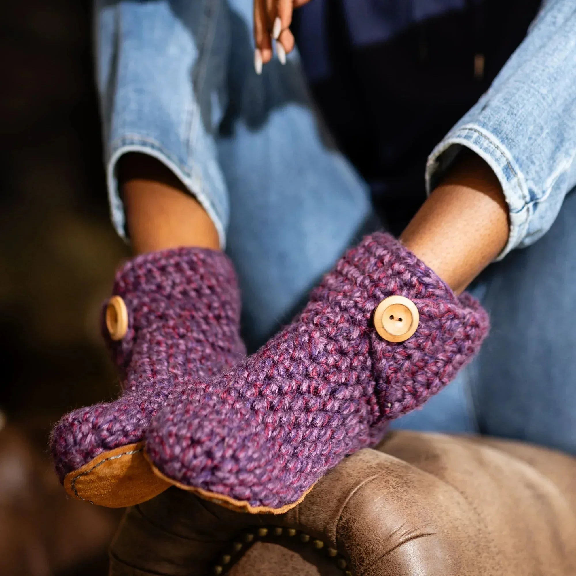 A close-up of a person wearing chunky hand-crocheted slipper boots in a heathered purple and pink colorway, featuring light brown wooden buttons and tan suede soles, paired with blue jeans.