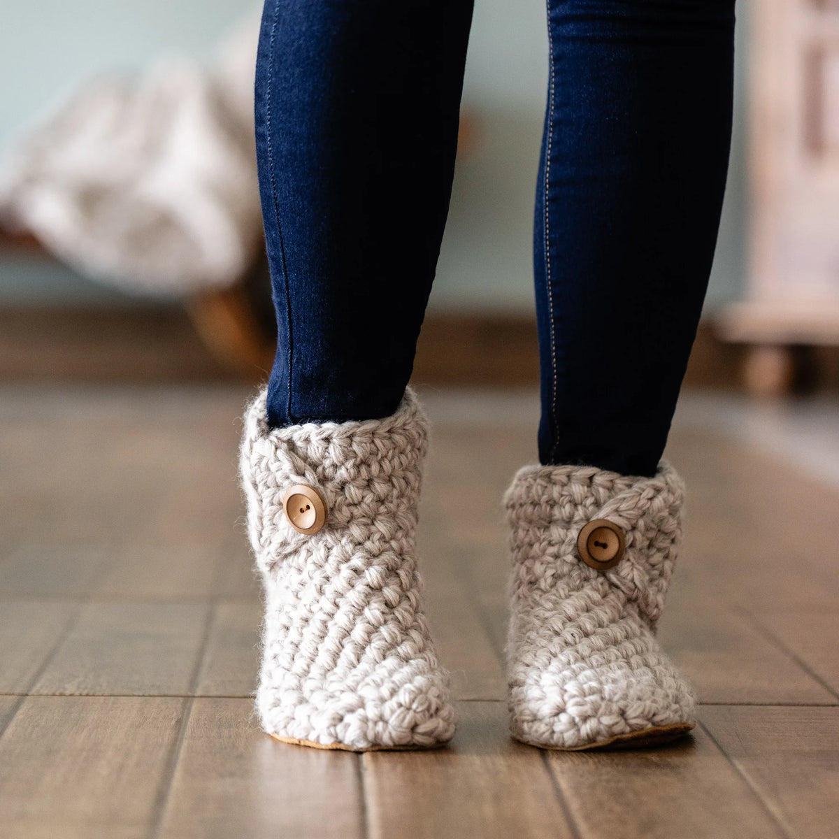 A woman wearing chunky cream-colored knit booties with wooden buttons, paired with dark blue skinny jeans, standing on a wooden floor.