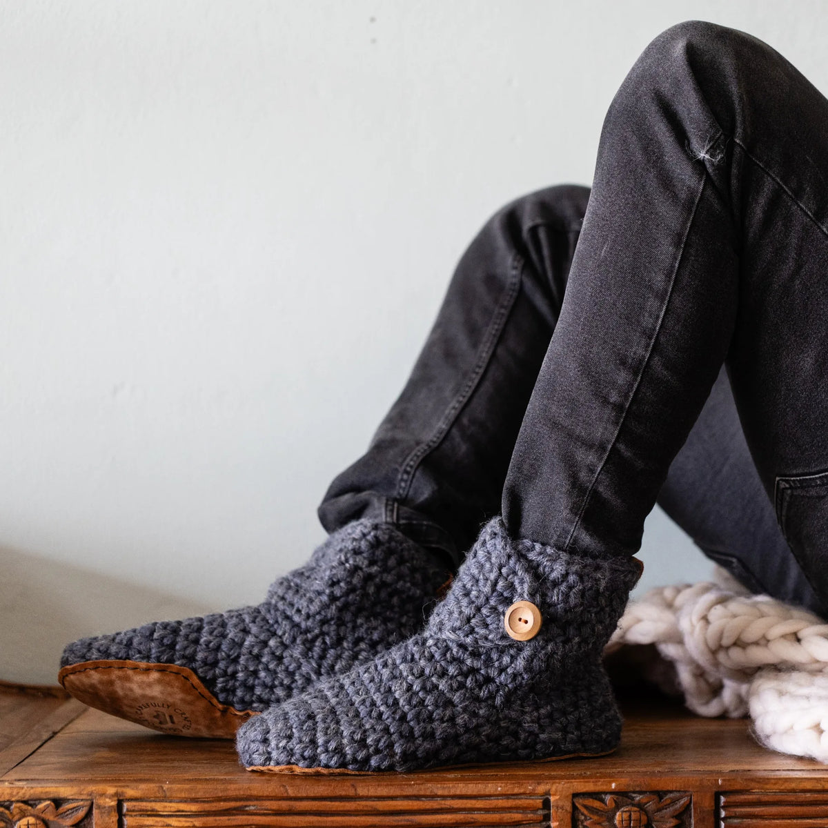 A man sitting on a wooden bench wearing dark charcoal gray jeans and chunky knit slippers in a dark gray color, with a small wooden button detail on the side.
