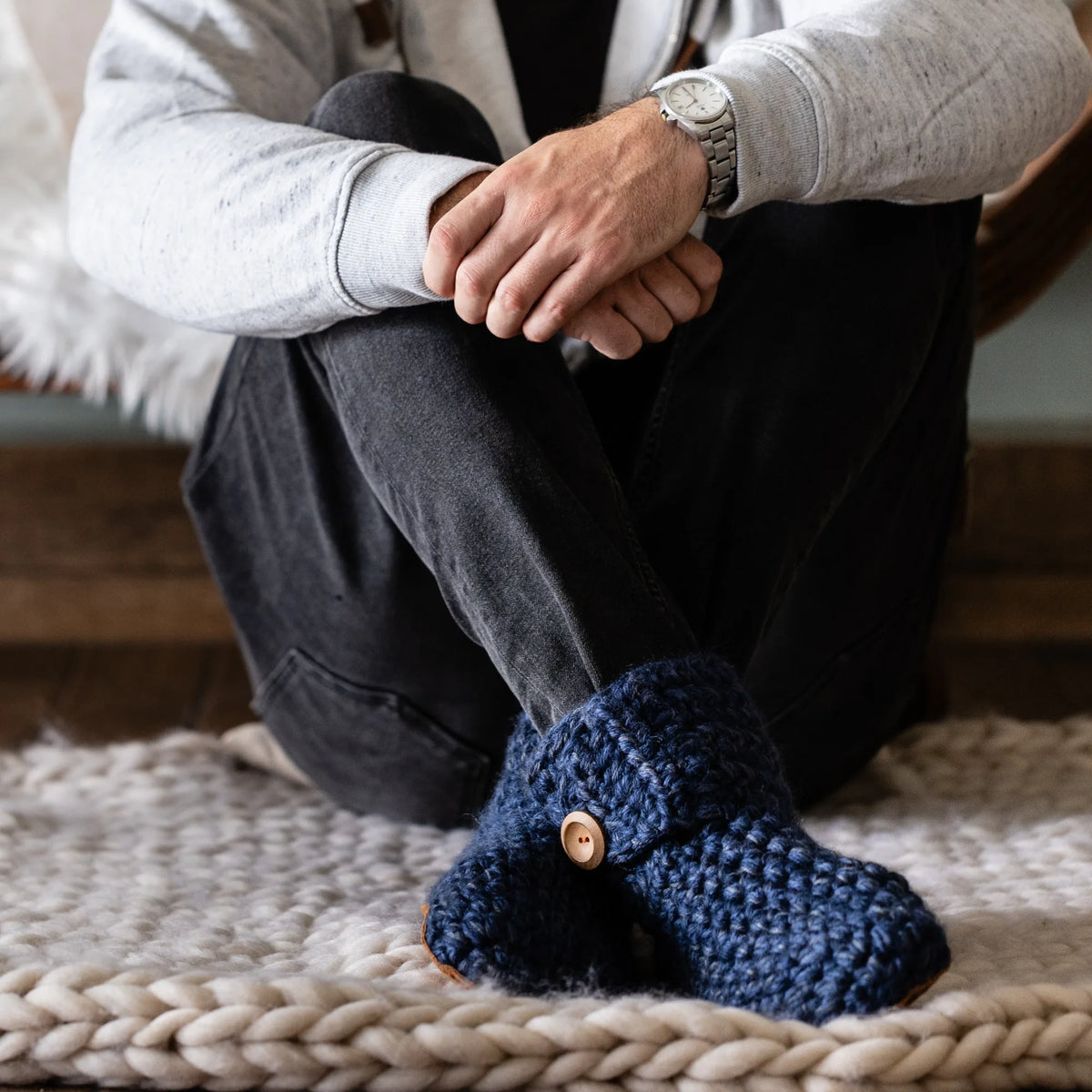 A man sitting cross-legged on a chunky knit rug wearing dark blue crocheted slippers with wooden buttons, black jeans, and a light gray sweatshirt.