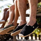 Two pairs of feet wearing black espadrilles with jute sole, sitting on a wooden bench outdoors.