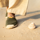 Man wearing khaki green espadrilles on a sandy beach with a shell nearby.