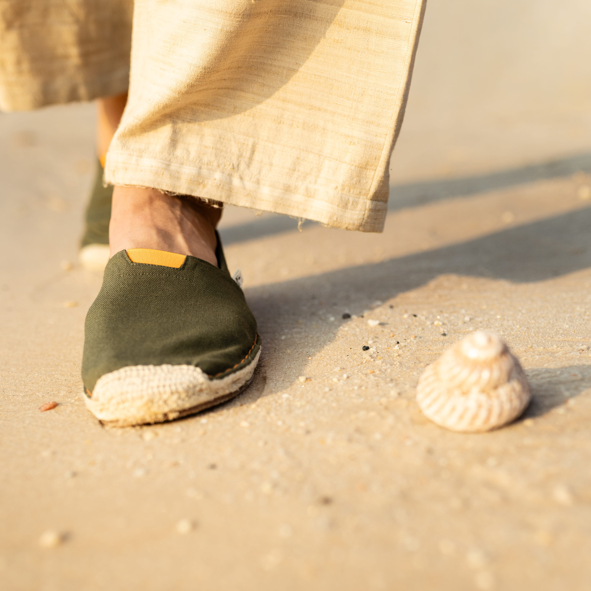 Man wearing khaki green espadrilles on a sandy beach with a shell nearby.
