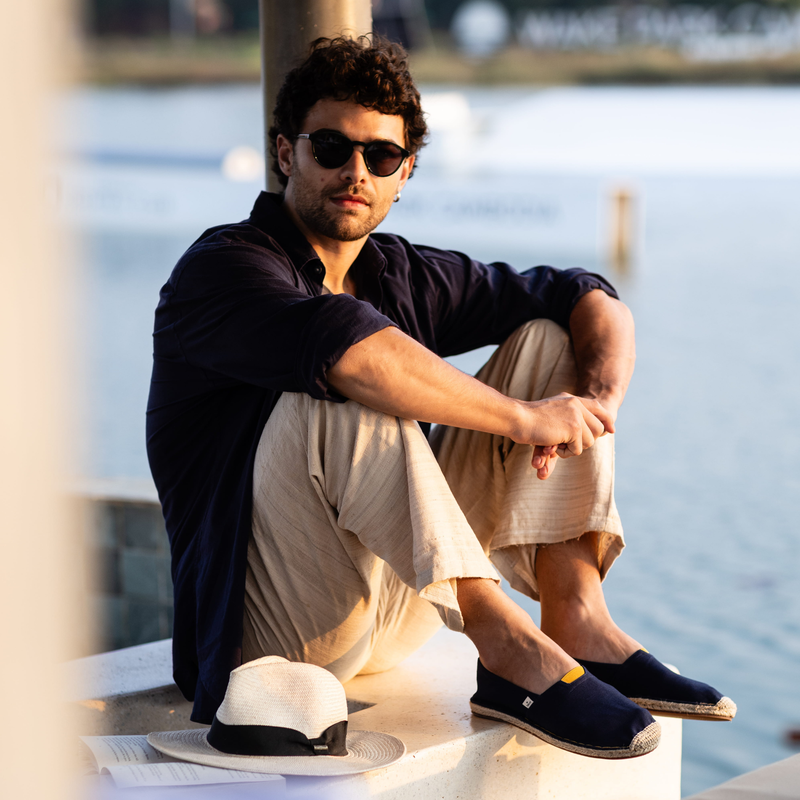 Man sitting by a waterfront wearing sunglasses, casual clothing and summer espadrilles