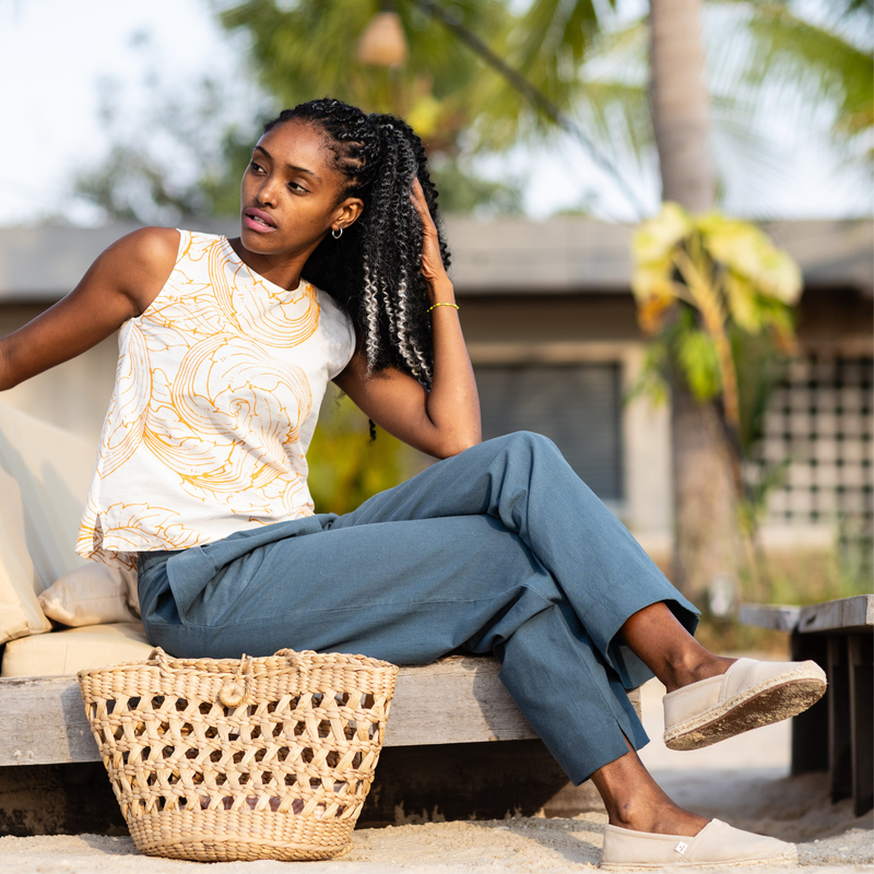 Woman sitting on a bench with a woven bag, surrounded by greenery wearing espadrilles