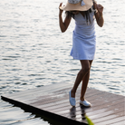 Woman in a light blue dress and sun hat standing on a wooden dock by the water.