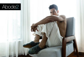 Man sitting on a chair wearing white pants and green slippers in a bright room.