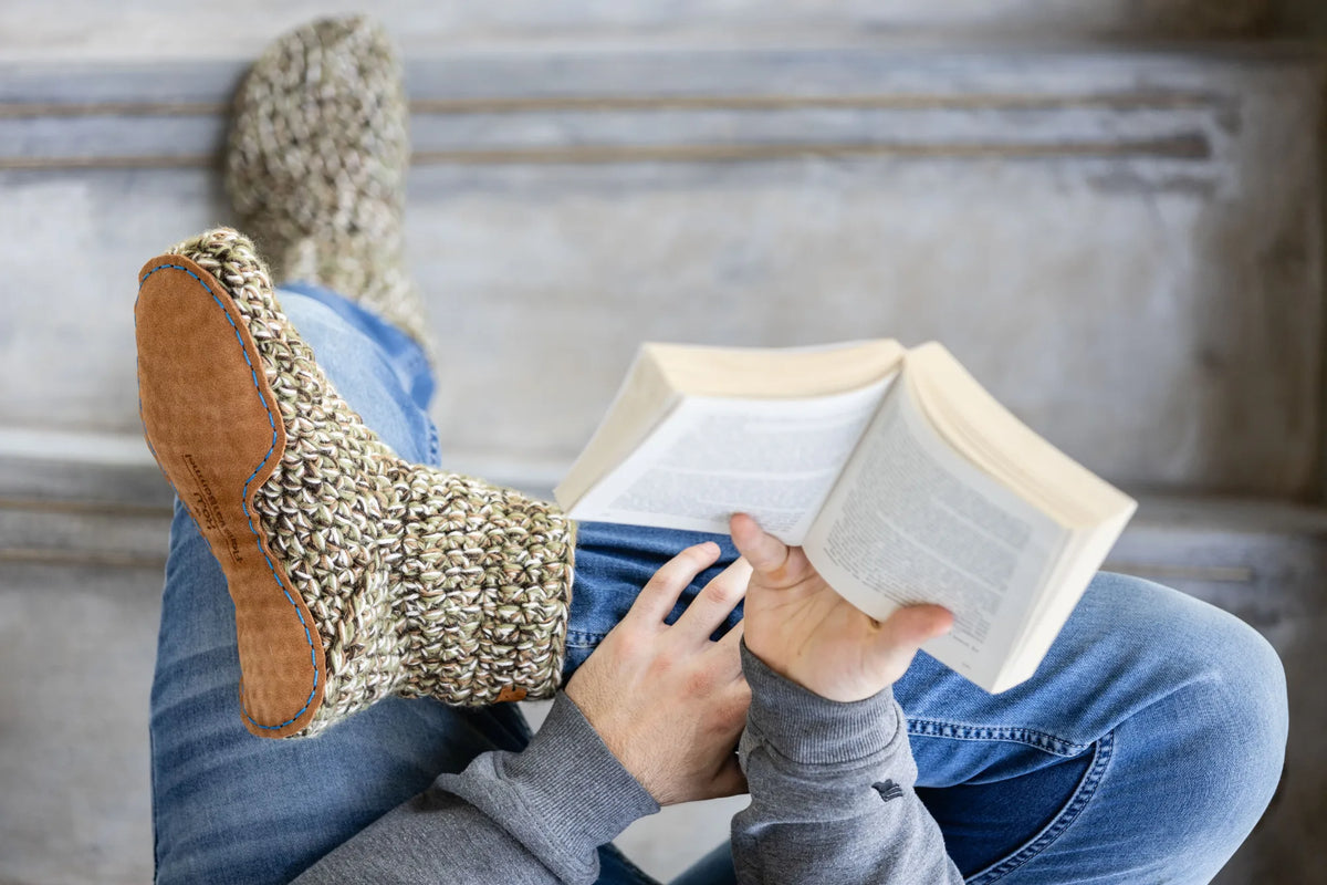 A person sitting on stone steps, holding an open book and wearing chunky knit slippers with leather soles and blue jeans.