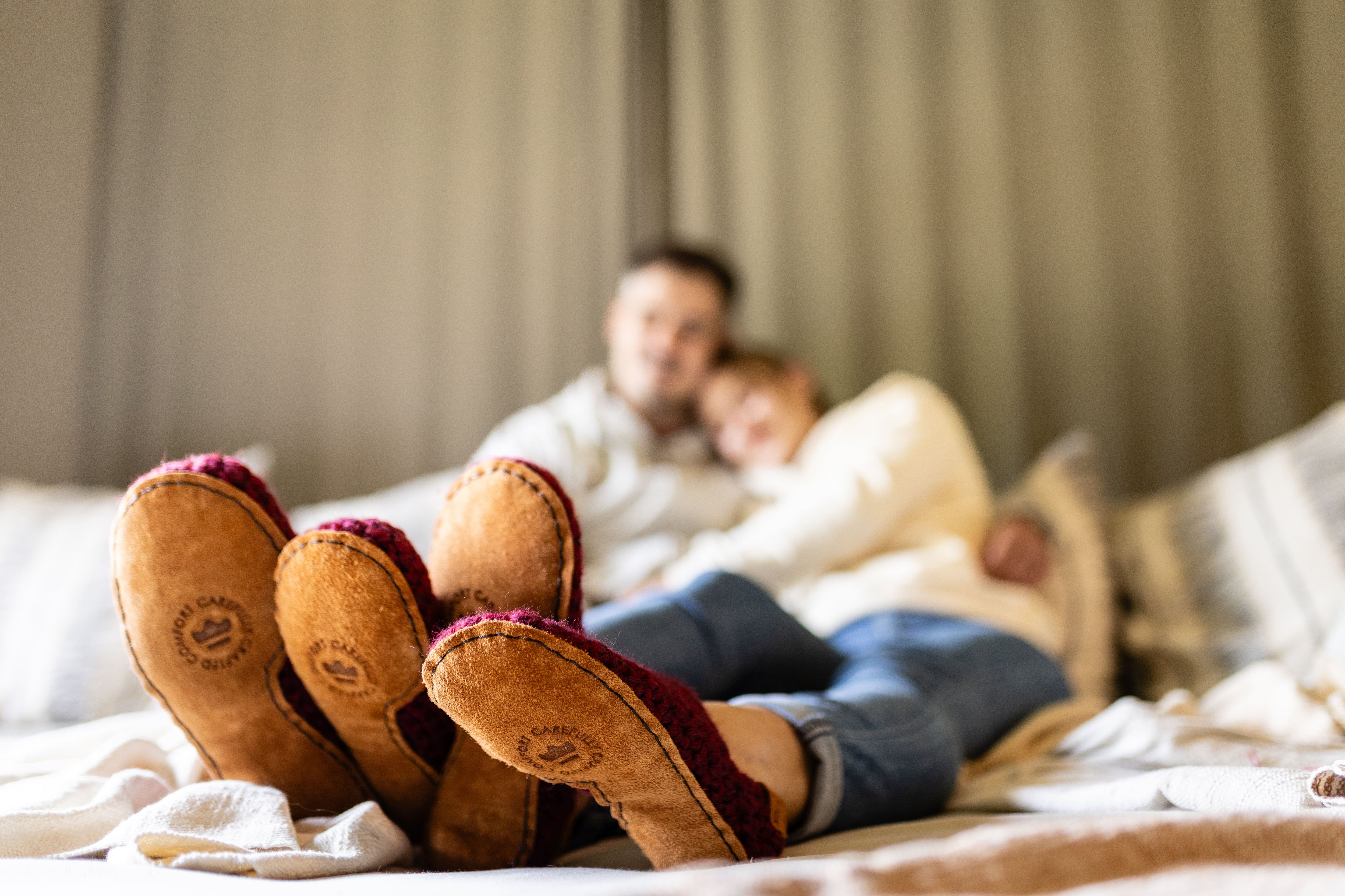Two pairs of feet in slippers with a blurred background of a couple lying on a bed.
