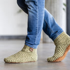 Green crocheted slipper boots with leather soles worn with blue jeans on a light wooden floor.