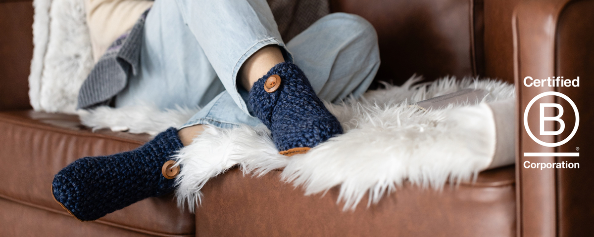 Woman sitting on couch wearing navy blue wool boot slippers with B Corp Certified logo