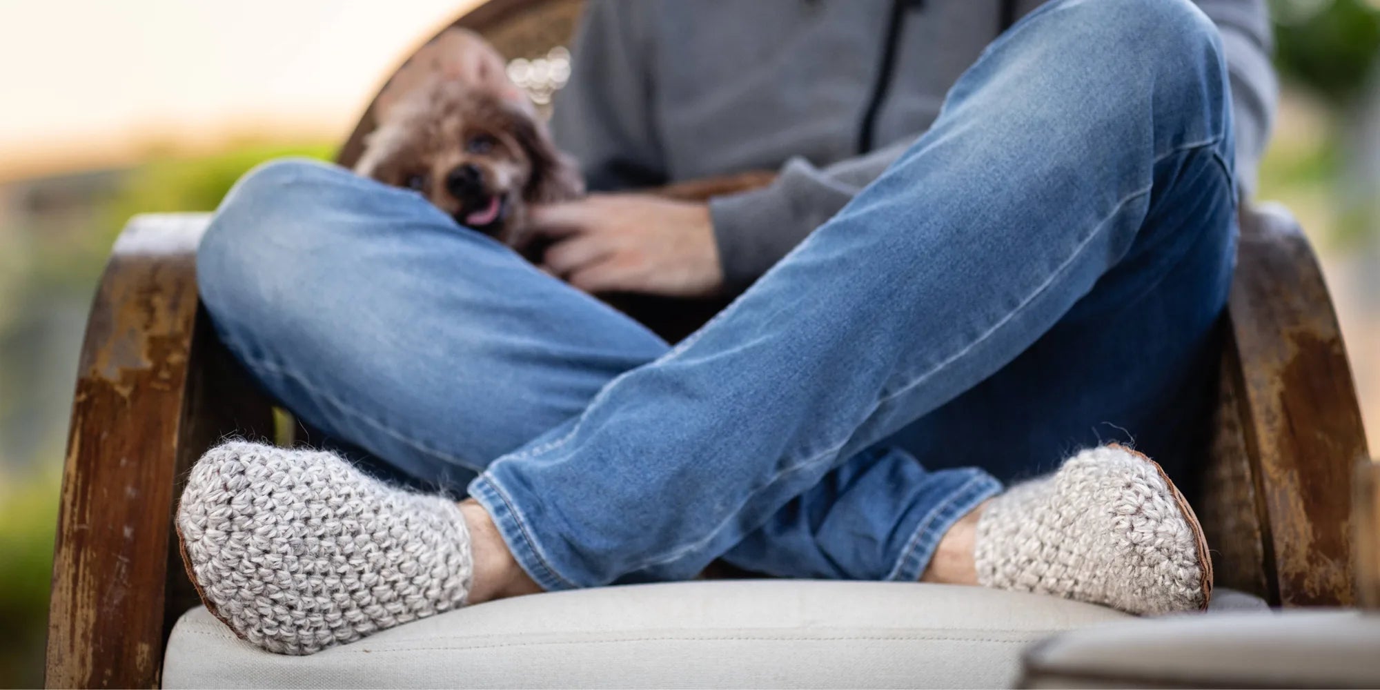 Person wearing blue jeans and off-white knit slippers sitting on a chair with a dog.
