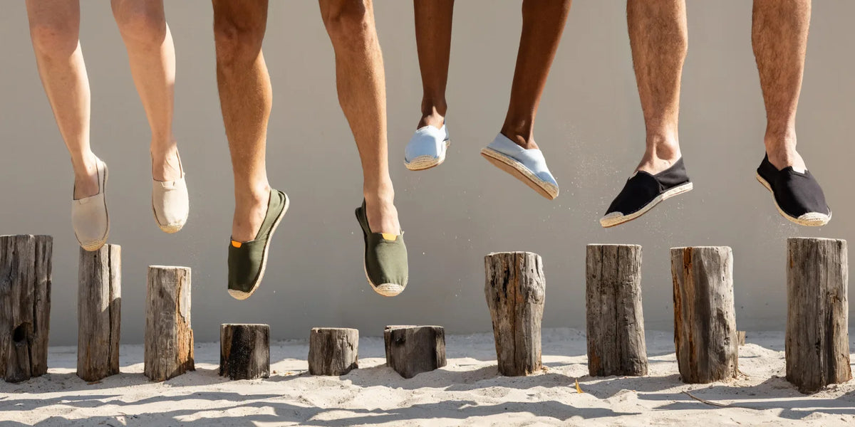 Several people's legs and feet are visible from the waist down, jumping over rustic wooden posts on sand. They are wearing casual slip-on espadrilles in beige, olive green, light blue, and black.
