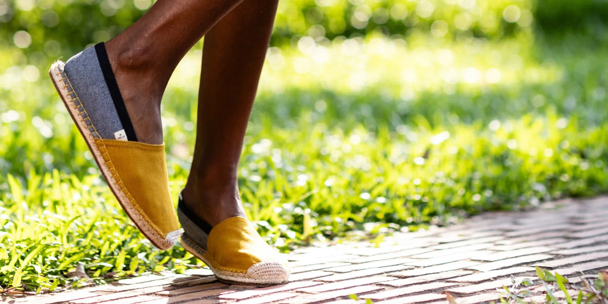A woman wearing yellow and gray espadrille slip-on espadrilles walking on a brick path outdoors with green grass in the background.