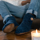 A person wearing cozy dark blue knit slipper socks with tan soles and a wooden button, sitting in jeans with a lit candle in the foreground on a wooden table.