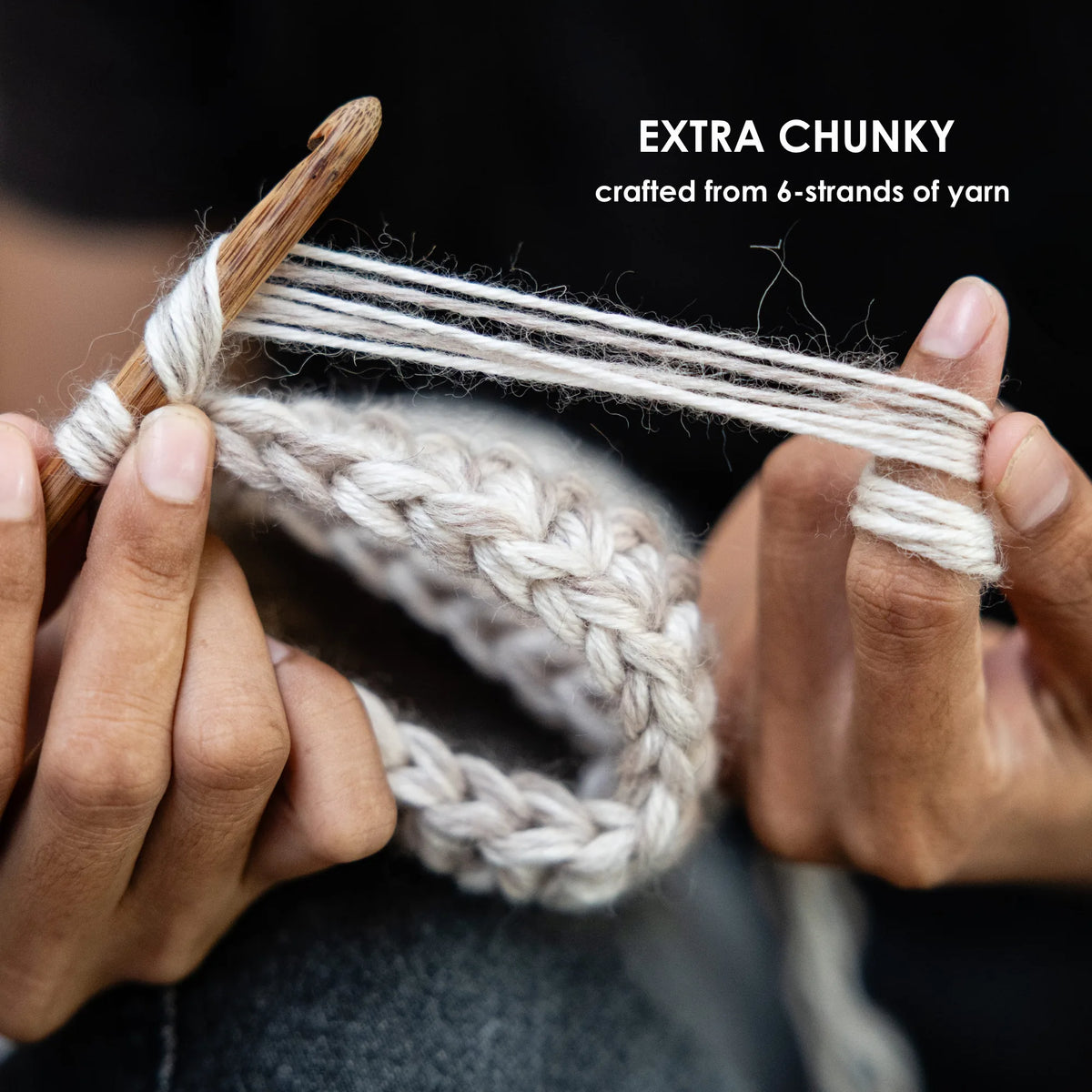 Close-up of hands knitting with chunky off-white yarn and a wooden crochet hook against a dark background, with text reading 'EXTRA CHUNKY crafted from 6-strands of yarn'.