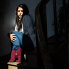 Woman sitting on a step wearing a white sweater, blue jeans, and red bootie wool slippers in a dimly lit setting.