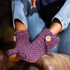 Lavender purple crocheted bootie slippers with wooden buttons worn by a person sitting on a brown chair