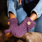 A close-up of a person wearing chunky hand-crocheted slipper boots in a heathered purple and pink colorway, featuring light brown wooden buttons and tan suede soles, paired with blue jeans.