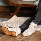 A man seated indoors wearing chunky cream knit slipper socks with tan leather soles and a small wooden button detail, paired with dark jeans. 