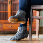 A woman wearing gray chunky crochet slipper boots by KOW with tan soles and wooden buttons, sitting on a wooden chair in a warm indoor setting. 
