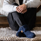 A man sitting cross-legged on a chunky knit rug wearing dark blue crocheted slippers with wooden buttons, black jeans, and a light gray sweatshirt.
